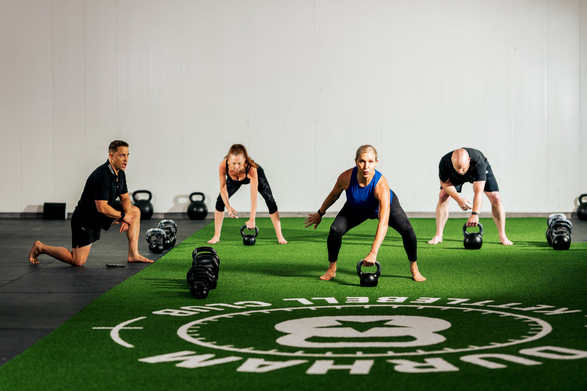A coach observes three adults practising kettlebell swings on green gym turf.